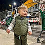 toddler, child, vest, pants, sneakers, sidewalk, night, streetlight, construction, fence, shopping_cart, urban, outdoor, happy, walking, person, clothing, smile, head, face