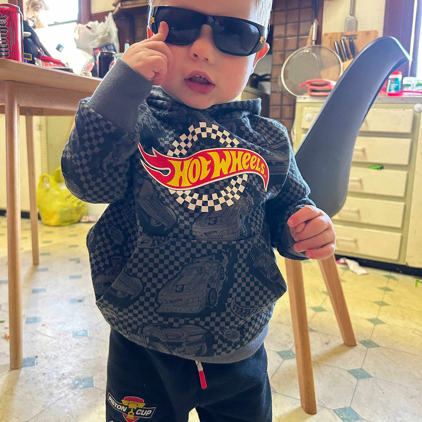 Brenn-Arthur is registered to the contest to win money with this photo: boy, cabinet, casual_clothing, chair, child, cute, expression, floor, hoodie, hot_wheels, indoor, kitchen, person, playful, standing, sunglasses, table, toddler, toy, young_child