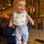 Logan is registered to the contest to win money with this photo: baby, child, smiling, indoor, table, adult, hand, balloon, celebration, lighting, wood, clothing, bib, socks, face, person, restaurant, decor, forest, happy