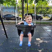 Logan is registered to the contest to win money with this photo: toddler, child, swing, playground, outdoor, park, smiling, curly_hair, black_swing, fence, tree, car, sidewalk, daylight, happy, seat, clothing, sock, person, fun