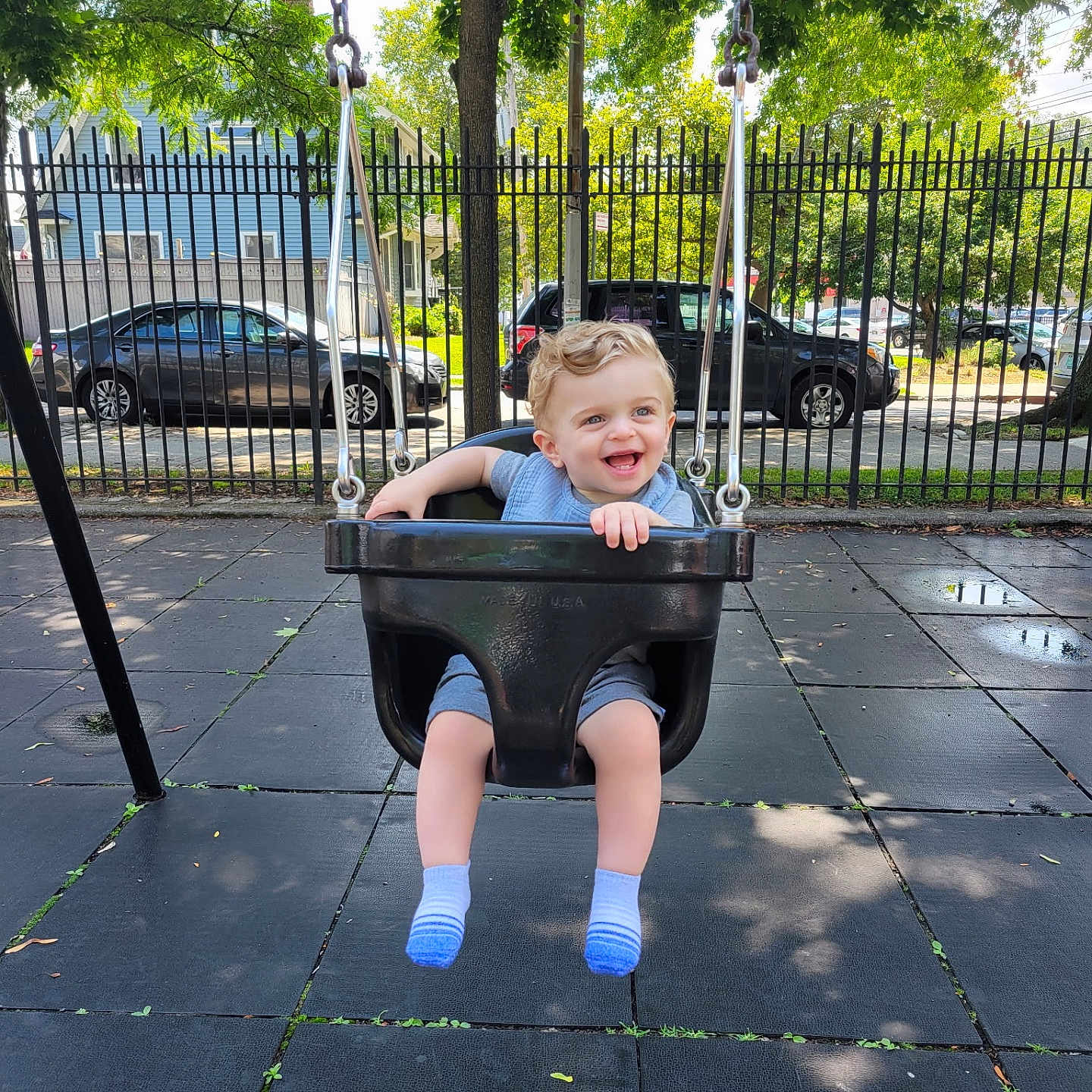 Logan is registered to the contest to win money with this photo: toddler, child, swing, playground, outdoor, park, smiling, curly_hair, black_swing, fence, tree, car, sidewalk, daylight, happy, seat, clothing, sock, person, fun