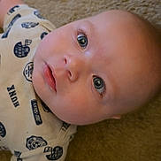 River is registered to the contest to win money with this photo: baby, infant, face, eyes, carpet, clothing, shirt, person, skin, head, child, lying_down, cute, portrait, expression, young, indoors, casual, looking_up, closeup