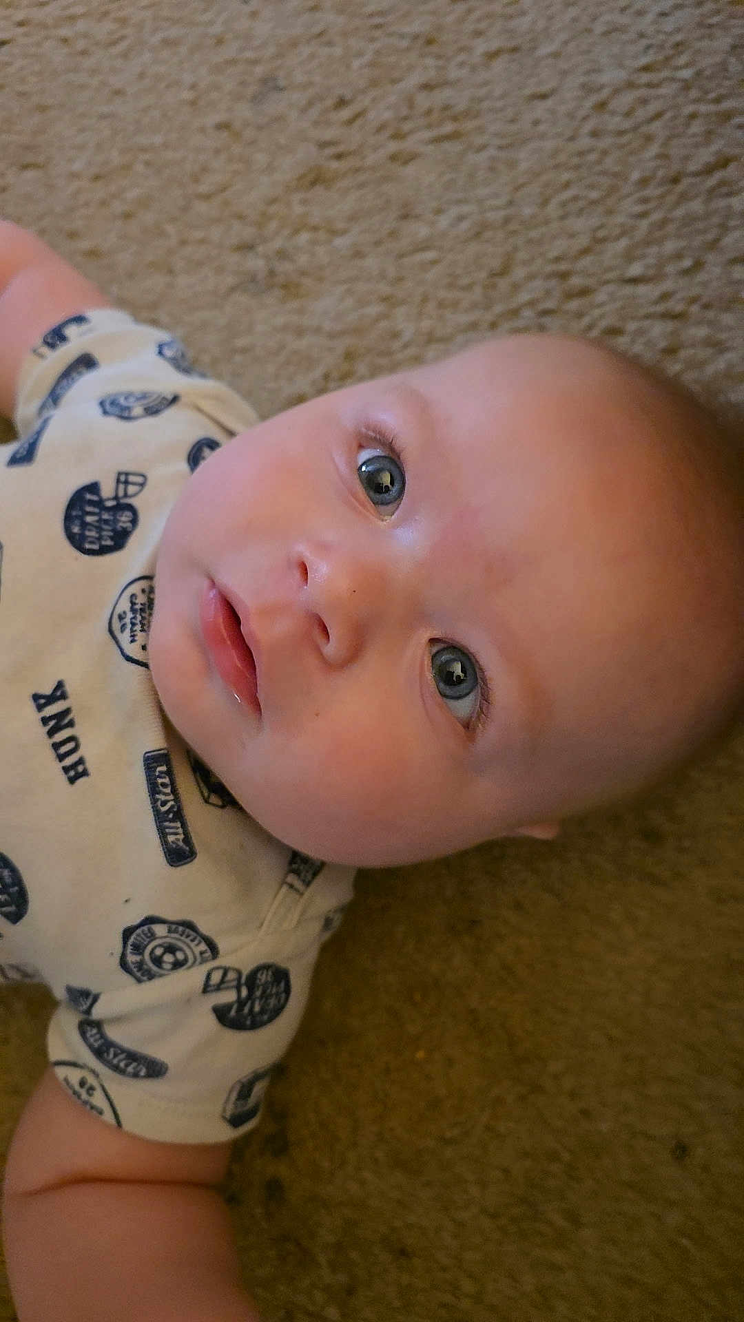 River is registered to the contest to win money with this photo: baby, infant, face, eyes, carpet, clothing, shirt, person, skin, head, child, lying_down, cute, portrait, expression, young, indoors, casual, looking_up, closeup
