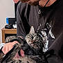 animal, cat, close_up, cozy, curious, face, facial_hair, gamer, gaming_console, holding, home, indoor, necklace, person, pet, tabby_cat, tattoo, tshirt, whiskers, wooden_table
