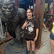 Ashley is registered to the contest to win money with this photo: child, statue, bigfoot, peace_sign, smile, tshirt, shorts, sneakers, bracelet, outdoor, brick_pavement, wooden_post, hand_gesture, person, sculpture, tourist_spot, daylight, happy, casual_clothing, fun