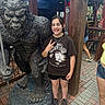 child, statue, bigfoot, peace_sign, smile, tshirt, shorts, sneakers, bracelet, outdoor, brick_pavement, wooden_post, hand_gesture, person, sculpture, tourist_spot, daylight, happy, casual_clothing, fun