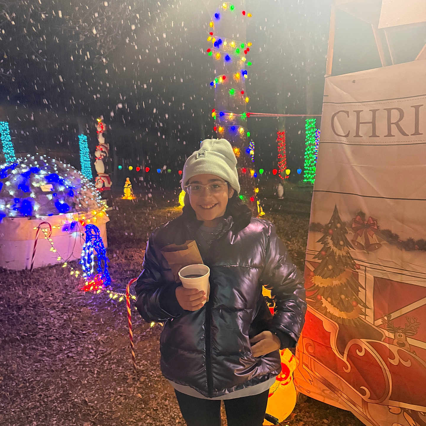 Ashley is registered to the contest to win money with this photo: child, smiling, winter_clothing, beanie, glasses, puffer_jacket, night, snow, holiday_lights, christmas_decorations, festive, outdoor, holding_cup, celebration, festive_banner, ground, colorful_lights, happy, person, event