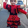 child, girl, dress, polka_dot, red_clothing, street, parked_car, portrait, smile, hair_clip, shoes, sidewalk, pavement, jeans, ruffles, outdoors, urban, buildings, playful, looking_at_camera