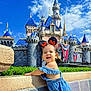 toddler, child, blue_romper, minnie_mouse_ears, castle, disney, stone_wall, greenery, blue_sky, clouds, happy, smile, outdoor, sunny, theme_park, architecture, tourist, holiday, playful, portrait