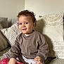 Oscar is registered to the contest to win money with this photo: toddler, child, curly_hair, toy, pink, hearts, couch, cushions, striped_pants, brown_sweater, indoor, smiling, seated, cozy, home, soft_lighting, portrait, cute, happy, casual_clothing