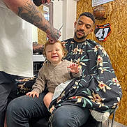 Oscar joined the competition — help win amazing prizes! toddler, man, barber, haircut, tattoos, barber_cape, chair, smile, hair, indoor, wood_paneling, sports_memorabilia, basketball, sign, casual_clothing, beard, people, child, happy, hairdresser