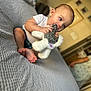 baby, child, stuffed_animal, toy, couch, gray, indoor, person, feet, white_shirt, blue_eyes, short_hair, blurred_background, home, floor, adult, walking, soft_toy, cute, seated
