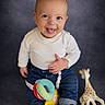 baby, black_sneakers, casual_clothing, child, cute, eyes, face, giraffe_toy, happy, infant, jeans, photography, plush_toy, portrait, sitting, smiling, studio_backdrop, tongue_out, toy_ring, white_shirt