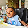 baby, child, high_chair, bib, spoon, food, messy, face, indoor, home, plant, furniture, cute, infant, feeding, expression, person, seat, wall_decor, kitchen