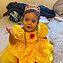 baby, child, dress, yellow_dress, crown, princess, bed, blanket, indoors, smiling, person, cute, happy, sitting, fabric, room, face, headwear, toddler, portrait