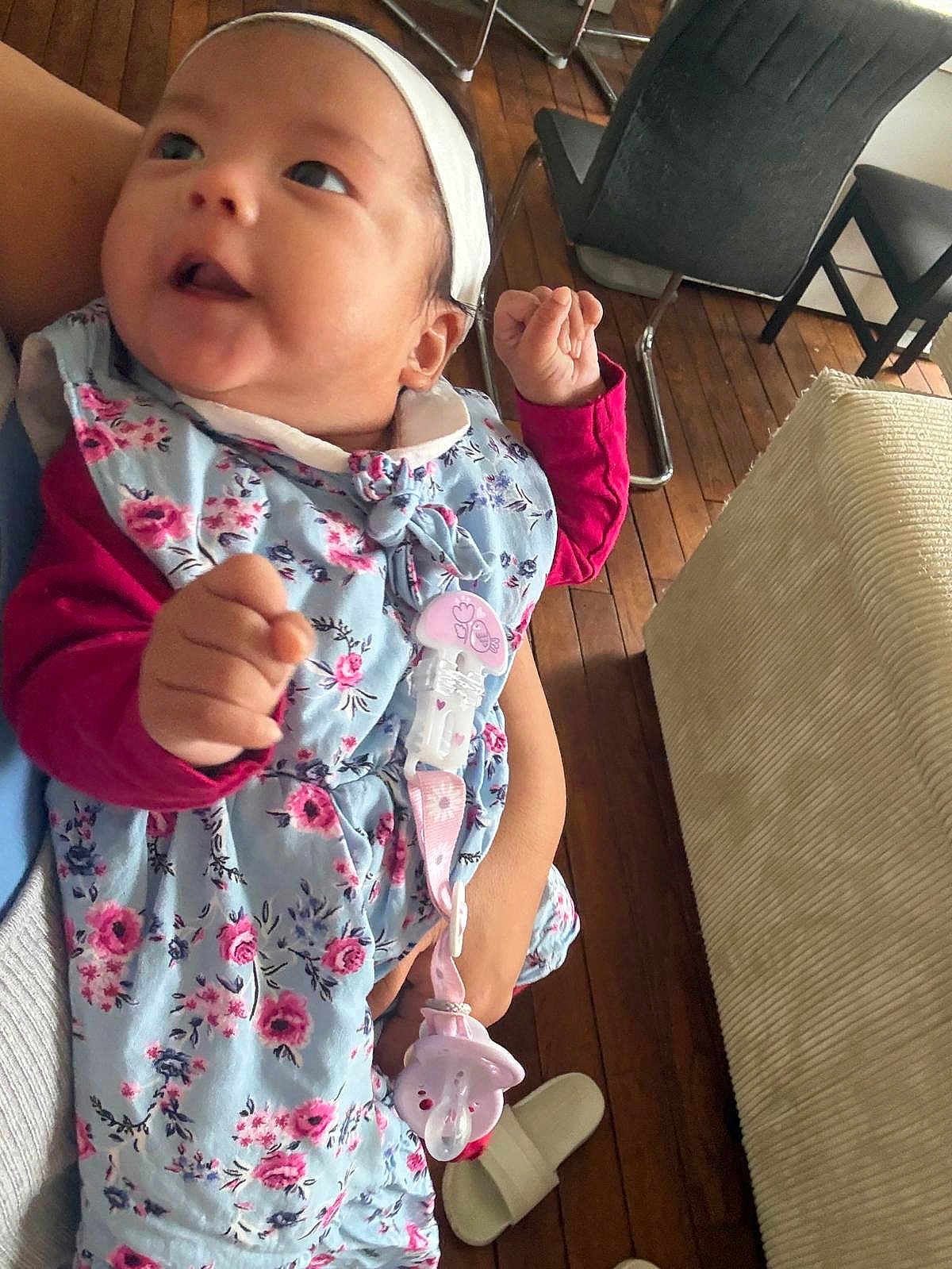 Kaylie a rejoint le concours — aidez-le/la à gagner de superbes lots ! baby, infant, child, face, headband, floral_dress, pacifier, pacifier_clip, hand, arm, cheek, smile, person, wooden_floor, chair, slipper, furniture, sitting, portrait, indoors