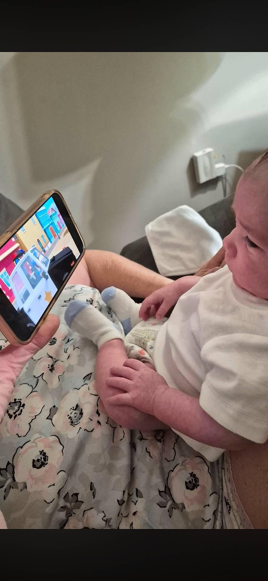 adult_arm, baby, blanket, cartoon, charging_cable, child, close_up, couch, diaper, floral_pattern, hand, indoor, infant, mobile_phone, onesie, phone_screen, portrait, seated, socks, wall_socket