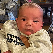Grayson joined the competition — help win amazing prizes! baby, newborn, infant, onesie, train_pattern, face, eyes, hand, holding, portrait, indoor, living_room, fan, metal_pitcher, table, wooden_floor, blanket, clothing, person, close_up