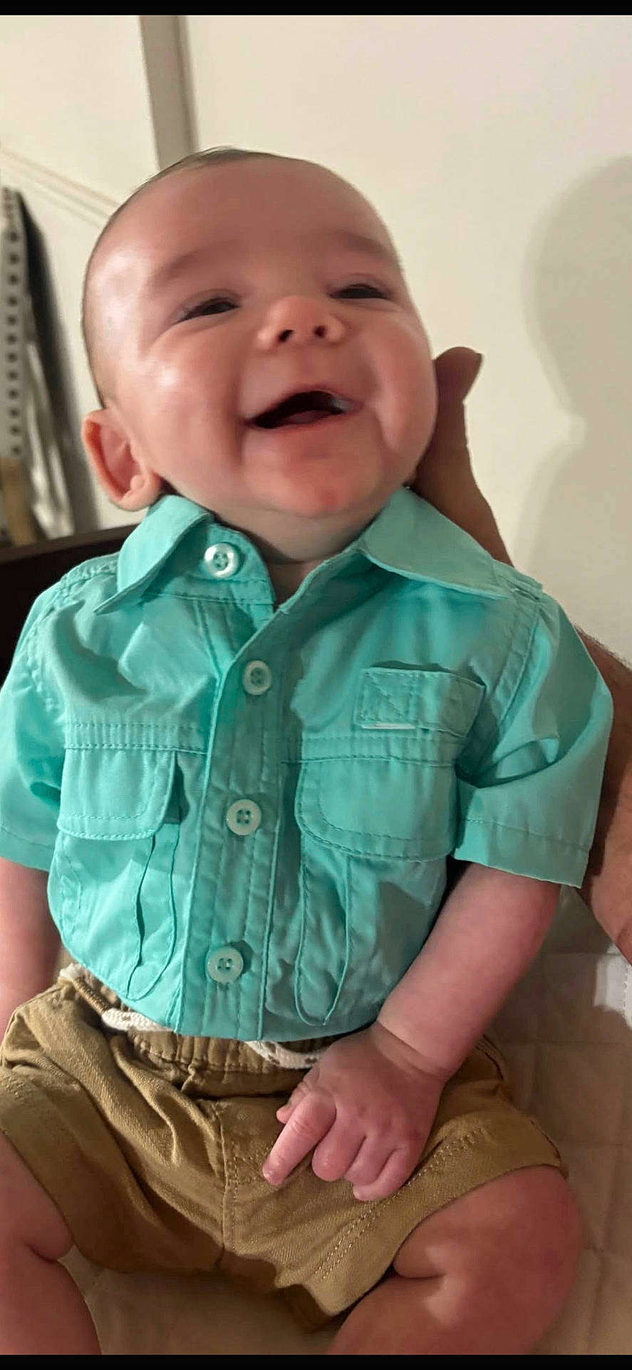 baby, baby_boy, background, casual_wear, child, clothing, cute, face, hand, happy, indoor, infant, khaki_shorts, person, portrait, sitting, smiling, support, turquoise_shirt, young_child