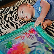 Grayson joined the competition — help win amazing prizes! baby, blue_clothing, child, colorful, curious, decor, face, floor, hand, indoor, infant, lying_down, pattern, person, play, soft_lighting, toy, water_mat, young, zebra_rug