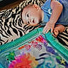 baby, blue_clothing, child, colorful, curious, decor, face, floor, hand, indoor, infant, lying_down, pattern, person, play, soft_lighting, toy, water_mat, young, zebra_rug