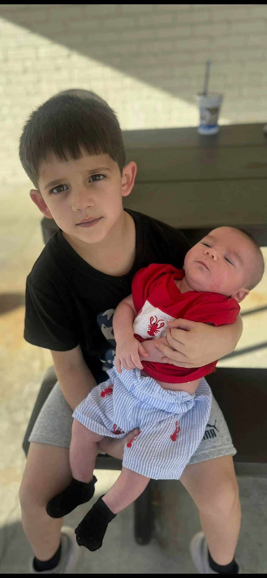 Alaiden joined the competition — help win amazing prizes! child, baby, siblings, holding, bench, picnic_table, outdoor, brick_wall, cup_with_straw, toddler, infant, portrait, red_shirt, striped_shorts, black_socks, shorts, serious_expression, sleepy_baby, hands, legs