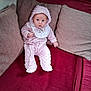 baby, infant, pink_clothing, hooded_outfit, bib, couch, cushions, red_fabric, indoor, person, seated, looking_at_camera, cozy, soft_texture, home_interior, child, cute, plush, small_hands, curious