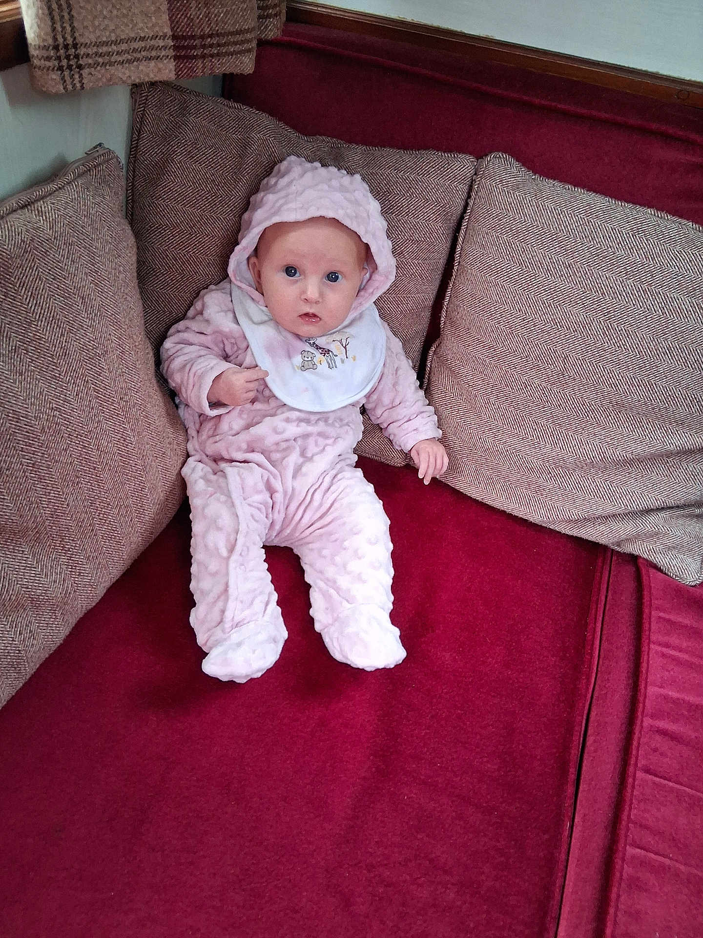 Kimberly joined the competition — help win amazing prizes! baby, infant, pink_clothing, hooded_outfit, bib, couch, cushions, red_fabric, indoor, person, seated, looking_at_camera, cozy, soft_texture, home_interior, child, cute, plush, small_hands, curious