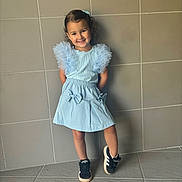 Deynah a rejoint le concours — aidez-le/la à gagner de superbes lots ! child, girl, smile, blue_dress, black_sneakers, curly_hair, standing, wall, tiles, playful, happy, posing, fashion, young, casual, footwear, indoor, person, cute, portrait