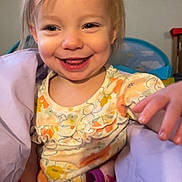 Caroline is registered to the contest to win money with this photo: toddler, child, smiling, happy, indoor, blanket, floral_clothing, face, hand, portrait, cute, baby, young_child, person, cozy, colorful, warm, expression, playful, hair