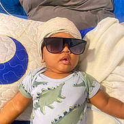Jace is registered to the contest to win money with this photo: baby, sunglasses, turban, dinosaur_pajamas, bed, pillow, blanket, child, cute, indoors, portrait, relaxing, fashion, apparel, person, sleepwear, cozy, infant, resting, soft_furnishing