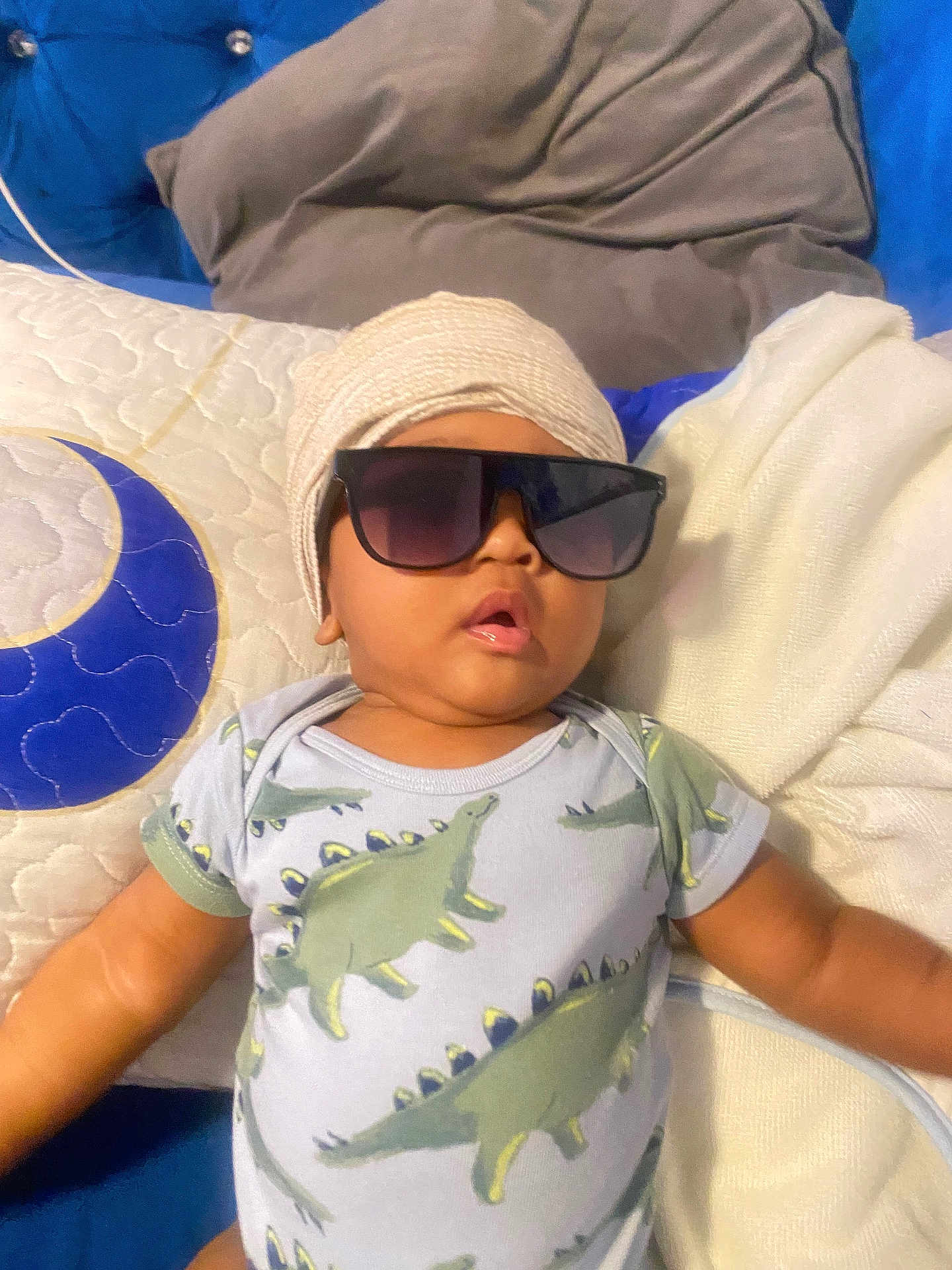 Jace is registered to the contest to win money with this photo: baby, sunglasses, turban, dinosaur_pajamas, bed, pillow, blanket, child, cute, indoors, portrait, relaxing, fashion, apparel, person, sleepwear, cozy, infant, resting, soft_furnishing