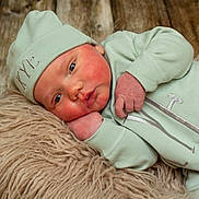 Kye is registered to the contest to win money with this photo: baby, infant, mint_green_clothing, hat, blue_eyes, fluffy_rug, wooden_floor, sleepwear, cute, newborn, soft_texture, portrait, lying_down, face, hand, skin, expression, cozy, indoors, closeup