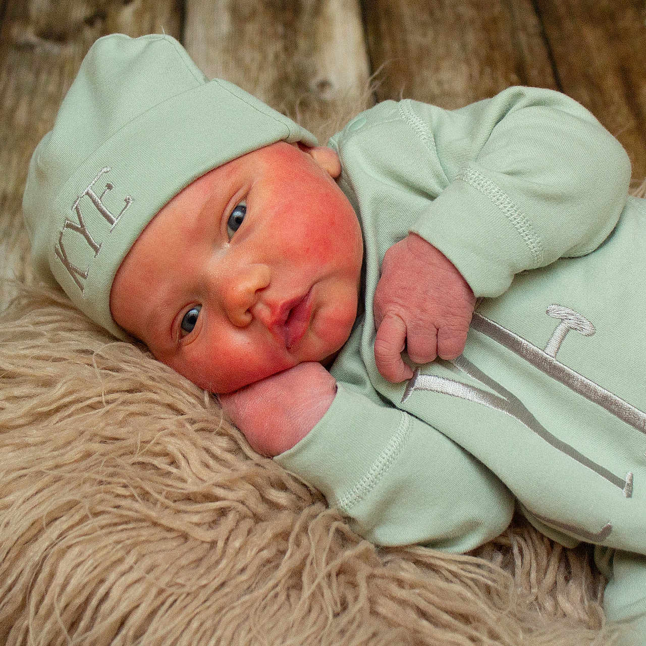 Kye is registered to the contest to win money with this photo: baby, blue_eyes, closeup, cozy, cute, expression, face, fluffy_rug, hand, hat, indoors, infant, lying_down, mint_green_clothing, newborn, portrait, skin, sleepwear, soft_texture, wooden_floor