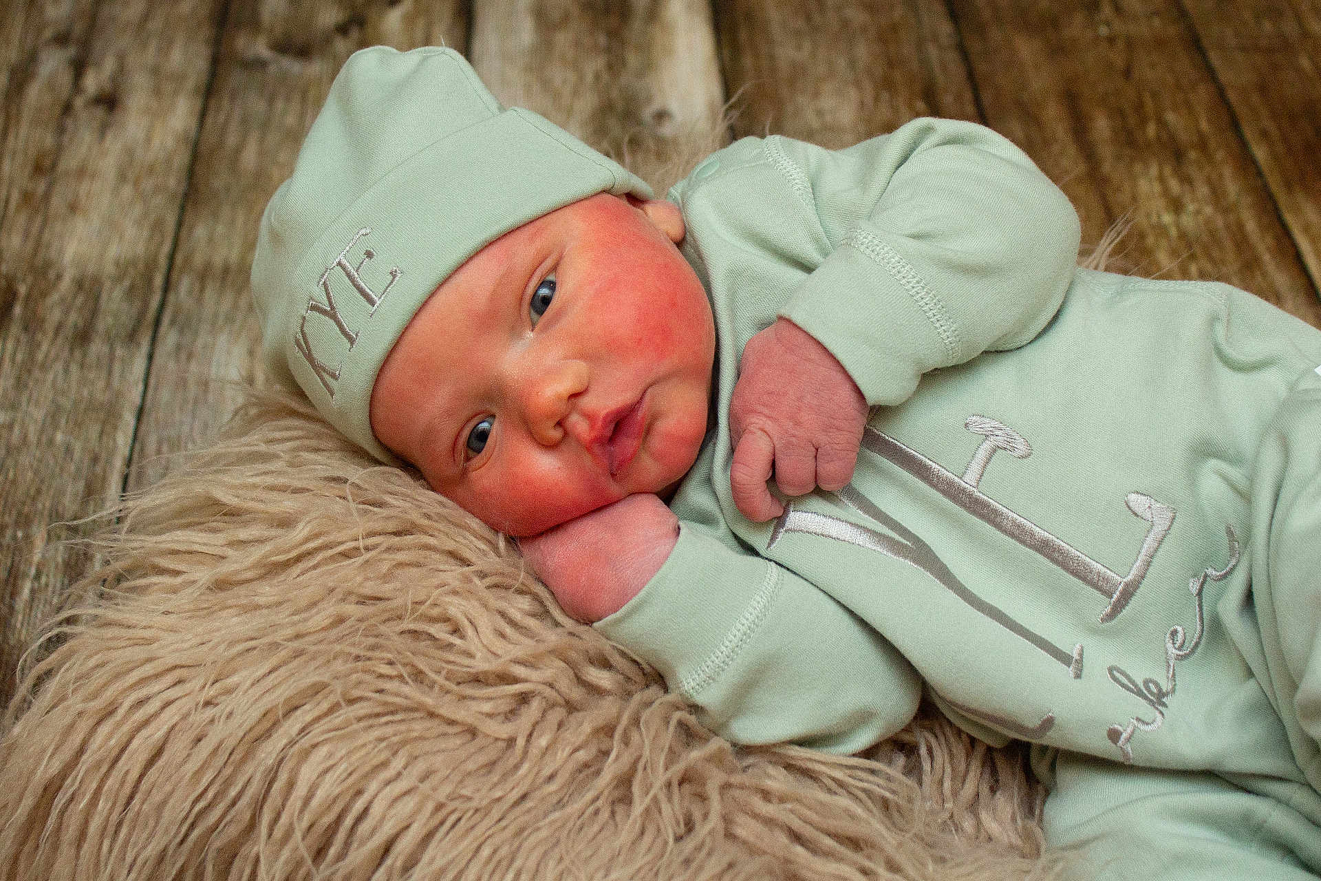 Kye is registered to the contest to win money with this photo: baby, infant, mint_green_clothing, hat, blue_eyes, fluffy_rug, wooden_floor, sleepwear, cute, newborn, soft_texture, portrait, lying_down, face, hand, skin, expression, cozy, indoors, closeup