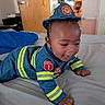 Jayson joined the competition — help win amazing prizes! baby, child, infant, firefighter_costume, costume, crawling, bed, bedroom, portrait, smiling, happy, playful, indoors, uniform, helmet, reflective_stripes, dark_skin, short_hair, furniture, photography