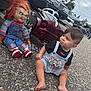 toddler, child, barefoot, toy_car, chucky_doll, pavement, outdoor, cars, vintage_cars, overcast_sky, playful, tongue_out, shorts, tshirt, person, red_hair, doll, toy, parking_lot, expression