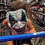 toddler, shopping_cart, store_aisle, mask, clown_mask, child, glowing, toy_store, aisle, retail, shopping, baby, seat, hand, foot, clothing, shelf, product, indoor, grocery