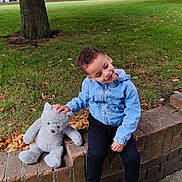 Malek is registered to the contest to win money with this photo: child, boy, teddy_bear, denim_jacket, black_pants, white_shoes, brick_wall, grass, tree, sidewalk, outdoor, smiling, playful, autumn_leaves, suburban, park, cute, young_child, casual_clothing, daylight