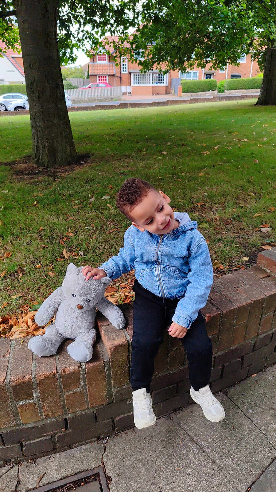 Malek is registered to the contest to win money with this photo: child, boy, teddy_bear, denim_jacket, black_pants, white_shoes, brick_wall, grass, tree, sidewalk, outdoor, smiling, playful, autumn_leaves, suburban, park, cute, young_child, casual_clothing, daylight