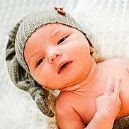 Jacob is registered to the contest to win money with this photo: baby, newborn, infant, child, hat, knitted_hat, button, hand, peace_sign, blanket, soft_texture, lying_down, close_up, portrait, skin, cute, expression, indoors, warm_colors, sleepwear