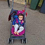 Aum is registered to the contest to win money with this photo: toddler, stroller, child, outdoor, pavement, jacket, pants, barefoot, purple, blue, greenery, trash_bins, curious, smile, seat_belt, pathway, wheel, person, casual, daylight