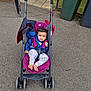 toddler, stroller, child, outdoor, pavement, jacket, pants, barefoot, purple, blue, greenery, trash_bins, curious, smile, seat_belt, pathway, wheel, person, casual, daylight