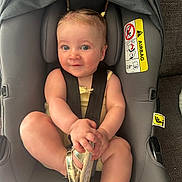 Holly is registered to the contest to win money with this photo: baby, car_seat, child, infant, seatbelt, gold_sandal, pigtails, cute, smile, indoors, soft_lighting, gray, fabric, straps, person, footwear, sitting, portrait, closeup, comfort