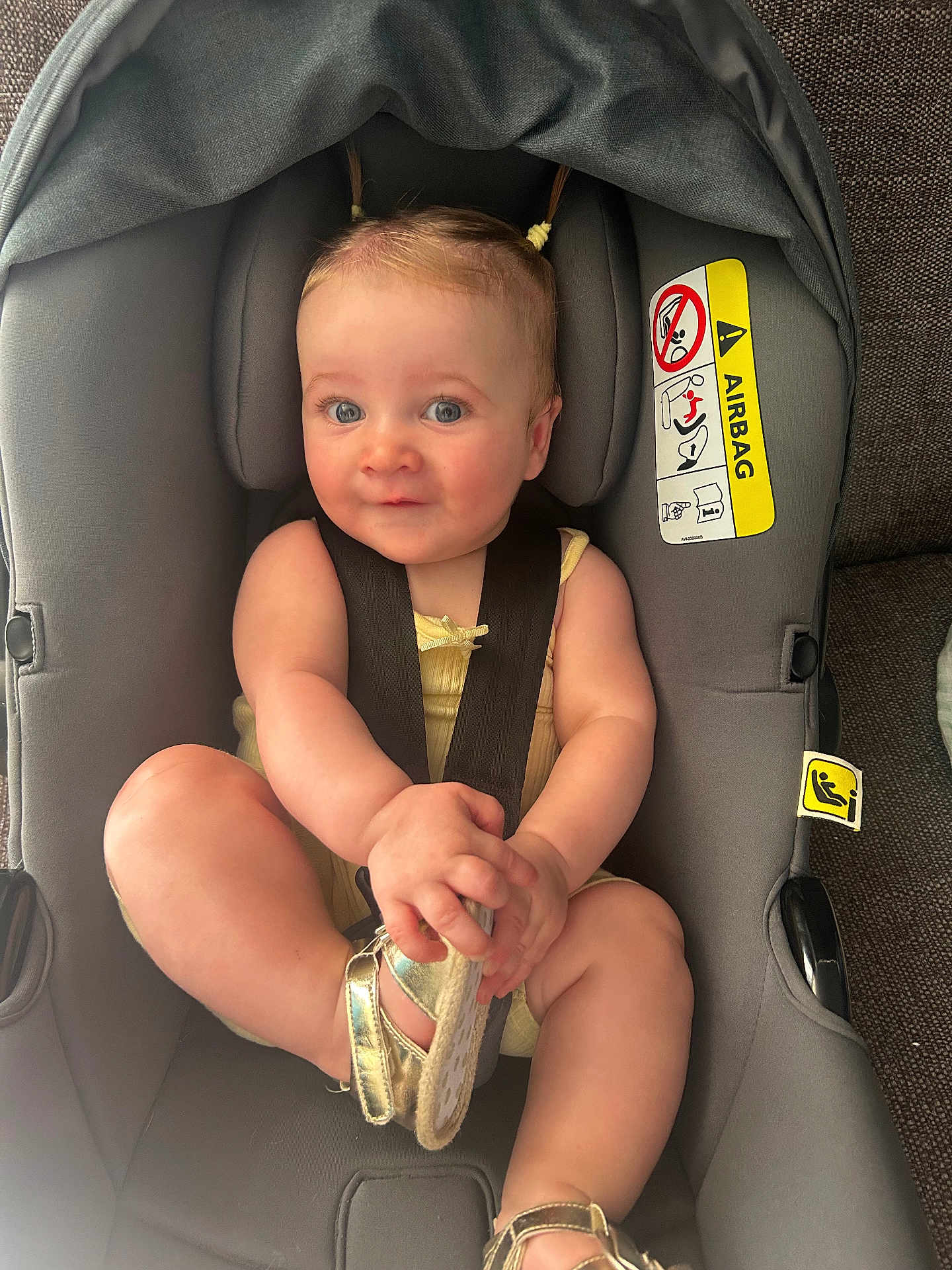 Holly is registered to the contest to win money with this photo: baby, car_seat, child, infant, seatbelt, gold_sandal, pigtails, cute, smile, indoors, soft_lighting, gray, fabric, straps, person, footwear, sitting, portrait, closeup, comfort
