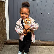 Nevaeh is registered to the contest to win money with this photo: child, toy, stuffed_animal, tiger, jacket, sneakers, door, smile, standing, outdoor, concrete, hair_bun, black_pants, playful, cute, person, young_child, happy, casual_clothing, urban