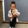 child, toy, stuffed_animal, tiger, jacket, sneakers, door, smile, standing, outdoor, concrete, hair_bun, black_pants, playful, cute, person, young_child, happy, casual_clothing, urban