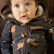 Jordan is registered to the contest to win money with this photo: baby, child, curly_hair, blue_eyes, onesie, animal_print, moose_pattern, cozy_clothing, indoor, couch, brown_background, cute, portrait, person, small_hands, seated, expression, curious, soft_fabric, hooded_clothing
