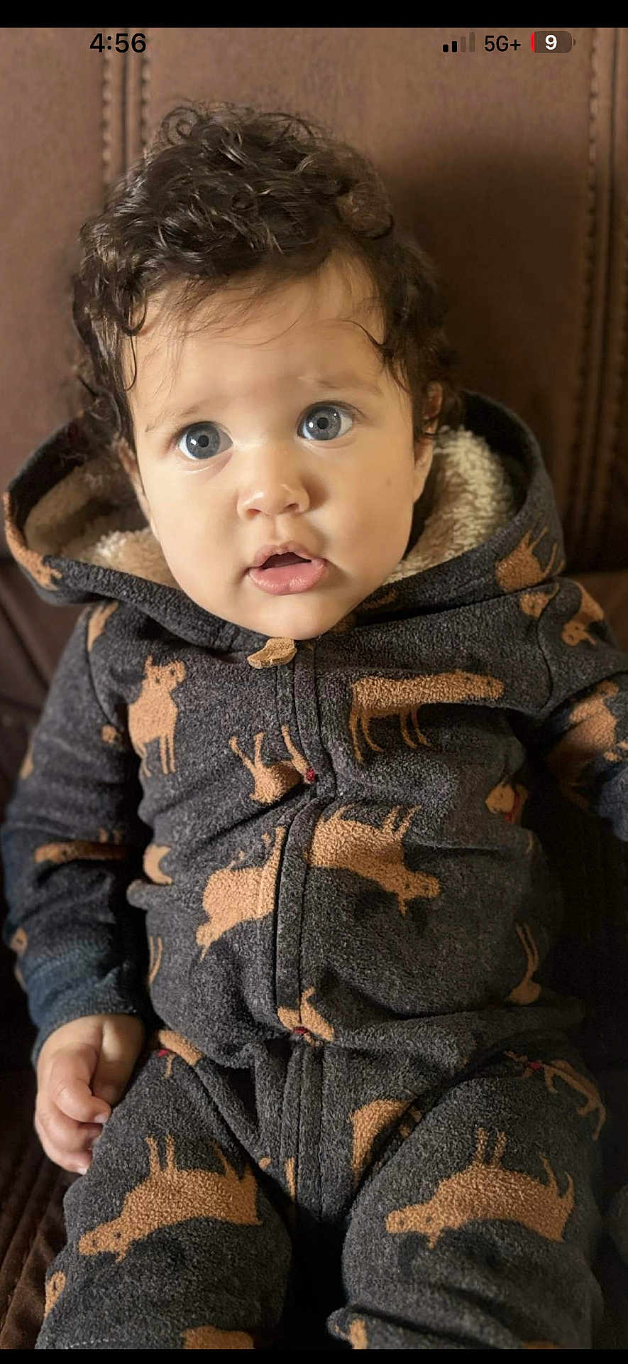 Jordan is registered to the contest to win money with this photo: baby, child, curly_hair, blue_eyes, onesie, animal_print, moose_pattern, cozy_clothing, indoor, couch, brown_background, cute, portrait, person, small_hands, seated, expression, curious, soft_fabric, hooded_clothing