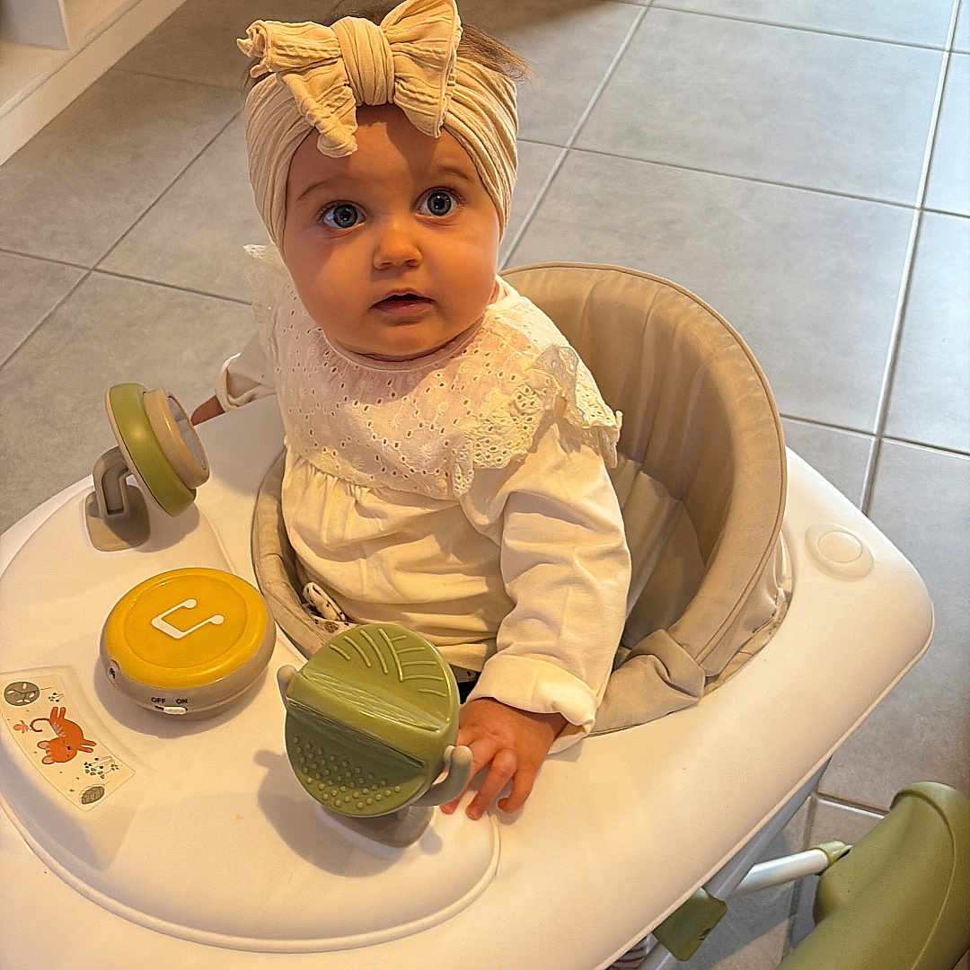 Tysaée participe au concours pour gagner de l'argent avec cette photo : baby, bow, child, clothing, curious, cute, floor, green, hand, headband, indoor, infant, looking_up, person, portrait, seat, tile, toy, walker, white